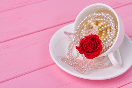 White cup full of pearl necklaces and red flower. Pink wooden desk.の写真素材