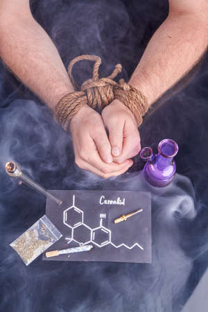 Slave of cannabis concept. Bound male hands with bong and smoking pipe. Molecule of cannabiol. Top view vertical shot.の写真素材