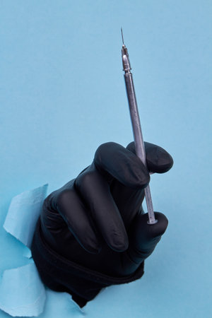 Hand holding gray cosmetic syringe on blue background. Black rubber latex glove.の写真素材