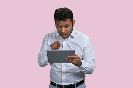 Shocked indian man pointing at someone on the tablet pc. Isolated on pink background.