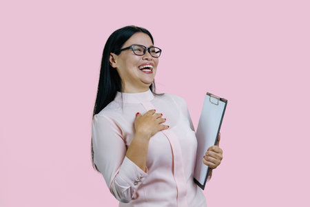 Young asian business woman in glasses holding clipboard and laughing very hard. Isolated on pink background.の写真素材