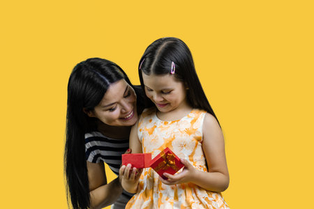 Little girl is opening a red gift box together with her mom. Isolated on yellow background.の写真素材