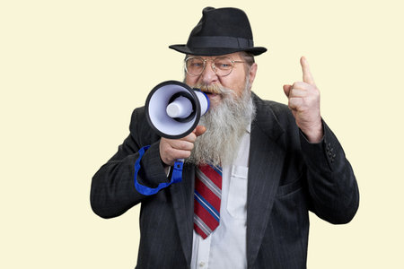 Portrait of aged senior businessman giving a speech in megaphone. Pastel yellow background.の写真素材