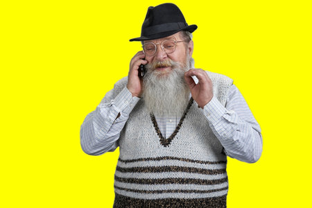 Portrait of bearded senior man with hat and beard talking on phone. Isolated on yellow background.