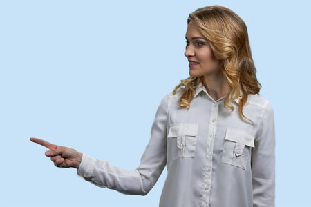 Young blonde woman pointing with her finger to the left. Pastel blue background.の写真素材