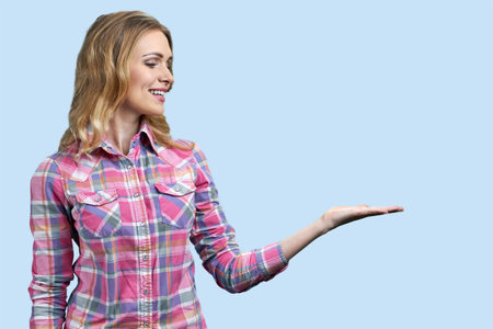 Smiling young blond woman is presenting copy space with one hand. Isolated on pastel blue background.の写真素材