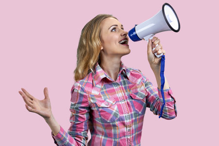 Portrait of young blonde blue-eyed woman speaking in megaphone. Attractive female with loudspeaker isolated on pink background.の写真素材