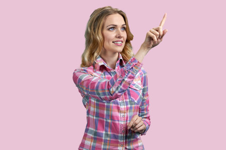 Portrait of blond woman pointing at transparent virtual screen. Isolated on pastel pink background.の写真素材