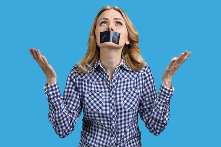 Portrait of a young woman with taped mouth is looking up. Attractive lady with her hands up. Isolated on vivid blue.の写真素材