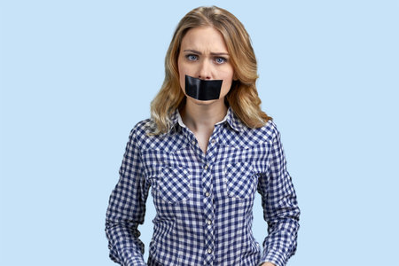 Portrait of angry stressed blond woman with mouth taped. Pastel blue background.の写真素材
