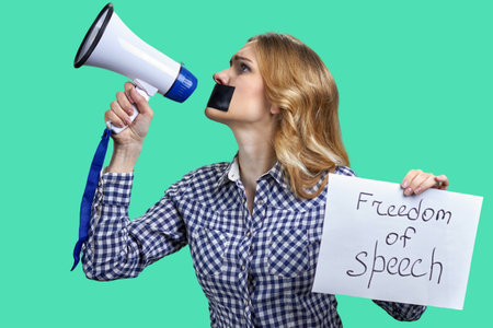 Young blonde woman holding megaphone and banner with freedom of speech inscription. Isolated on green background.の写真素材