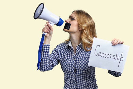 Young blonde woman with taped mouth holding megaphone and banner with censorship inscription. Isolated on yellow background.の写真素材