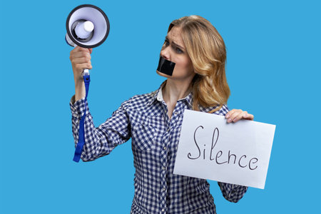 Blonde woman with taped mouth holding megaphone and banner with silence inscription. Isolated on vivid blue background.の写真素材