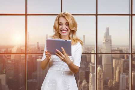 Smiling middle aged mature caucasian blonde lady holding digital tablet. Reading online social media news. Cityscape with evening sunset.の写真素材