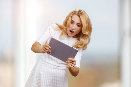 Excited expressive mature blonde woman holds digital tablet pc in both hands. Blurred indoors background.の写真素材