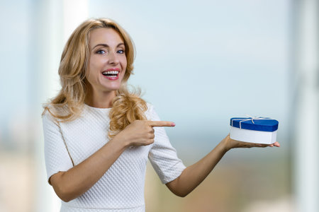 Positive middle aged blonde woman pointing at blue heart shape gift box on her hand. Abstract blurred indoor background.の写真素材