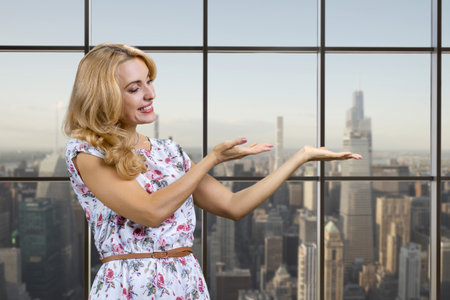 Sexy caucasian blonde woman shows copy place on her palm standing indoors. Checkered window with cityscape view in the background.の写真素材