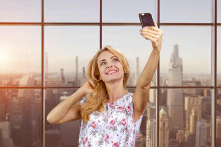 Smiling business woman making selfie photo on smartphone. Checkered window with cityscape in the background.の写真素材