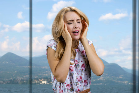 Young lady with long blond hair touching her head because of headache. Window with mountain resort in the background.の写真素材