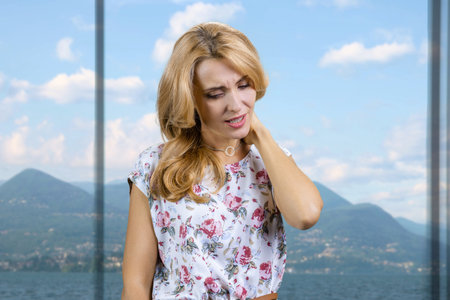Young middle aged blonde lady touching her neck because of pain. Blurred window with mountain landscape in the background.の写真素材
