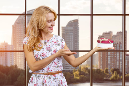 Blonde woman pointing at heart shape gift box with index finger. Windows with cityscape view in the background.の写真素材