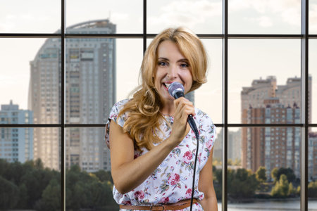 Blonde woman with microphone singing or giving a speech. Indoor window in the background.の写真素材