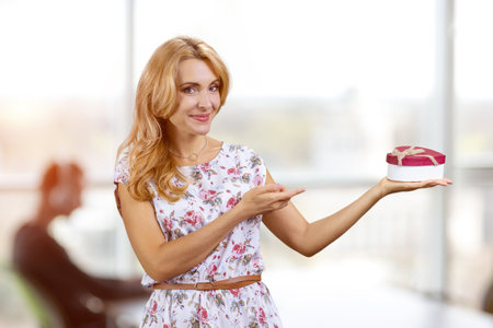 Blonde woman pointing at heart shape gift box. Office environment in the background.の写真素材