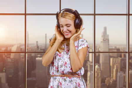 Portrait of happy blonde middle aged woman listening to music in headphones with closed eyes. Blurred urban cityscape in the background.の写真素材