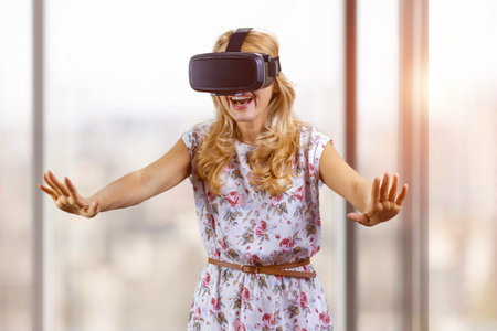 Amazed happy young woman experiences virtual reality stretch her arms. Indoor window in the background.の写真素材