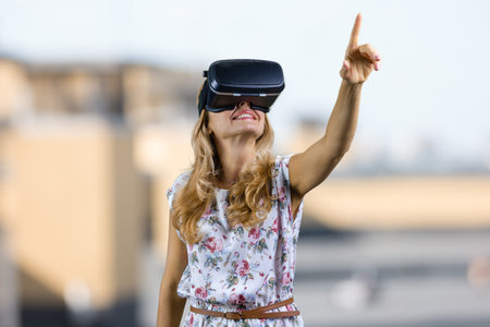 Amazed happy young woman wearing vr headset pointing up. Blurred outdoor background.の写真素材