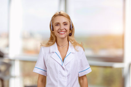 Smiling cheerful blonde woman in headset standing indoors. Female customer support operator. Blurred outdoor background.の写真素材