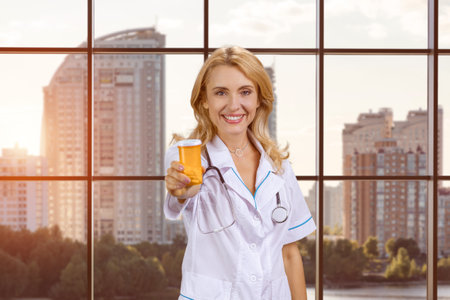 Portrait of a young female caucasian doctor giving you medicine bottle. Indoor window with cityscape view in the background.の写真素材