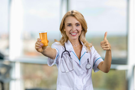 Happy blonde caucasian doctor gives you a medicine bottle and thumb up. Blurred window in the background.の写真素材