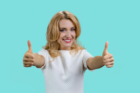 Portrait of a smiling blond woman gives both thumbs up. Isolated on turquoise.の写真素材