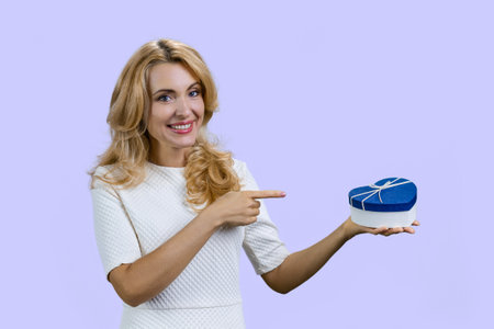 Happy blondie holding a blue heart shape gift box and pointing with index finger. Isolated on purple.の写真素材