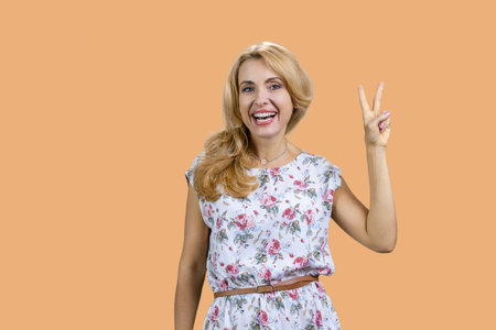 Portrait of attractive blonde mature woman shows victory gesture sign. Isolated on beige.の写真素材