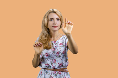 Portrait of a mature blonde woman taps something on an invisible imaginary virtual screen for copy space. Isolated on beige.の写真素材