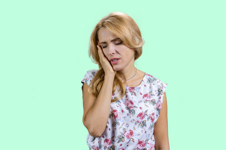 Portrait of mature blonde woman touching her cheek because of toothache. Dental illness on teeth. Isolated on turquoise.の写真素材