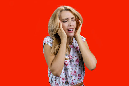 Portrait of mature blonde woman suffers from headache. Isolated on red.の写真素材