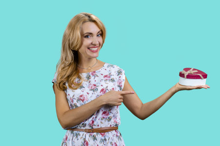 Blonde mature woman pointing at heart shape gift box. Isolated on turquoise.の写真素材