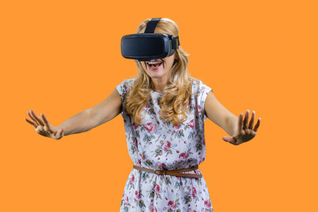 Happy shocked mature woman in virtual reality headset stretching her arms. Isolated on orange.の写真素材