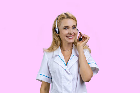 Portrait of cheerful blonde mature female doctor wearing headset. Isolated on pink.の写真素材