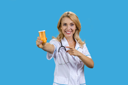 Portrait of happy blonde caucasian mature female doctor pointing at orange bottle of pills. Isolated on blue.の写真素材