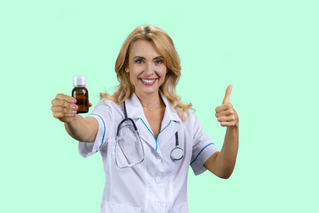 Portrait of happy caucasian female doctor holding a medicine bottle and gives her thumb up. Isolated on turquoise.の写真素材