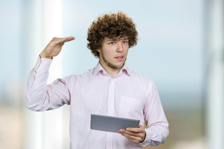 Portrait of a young man in white shirt holding a tablet pc showing height of something with hand. Male entrepreneur with curly hair giving a speech indoors.の写真素材