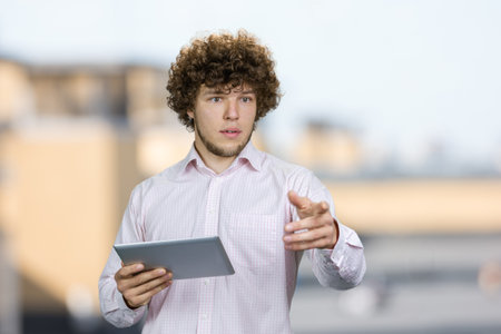 Portrait of a young man with curly hair holding a tablet pc and pointing at something. Blurred urban background.の写真素材