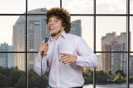 Happy excited young man with microphone standing indoors. Checkered window with residential area in the background.の写真素材