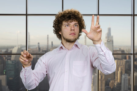 Portrait of a young man with curly hair and white shirt using invisible virtual screen for work. Indoor window background with cityscape view.の写真素材