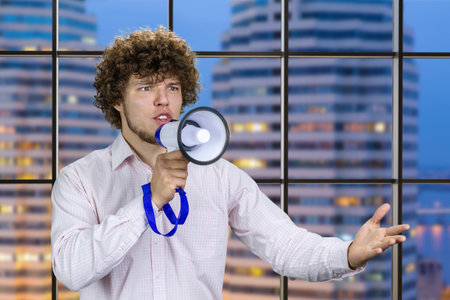 Handsome young man with curly hair in white shirt speaks in megaphone. Indoor window with night city view in the background.の写真素材