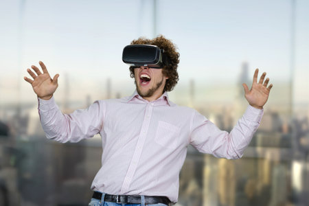 Portrait of young excited man with curly hair experiencing virtual reality. Blurred cityscape in the background.の写真素材
